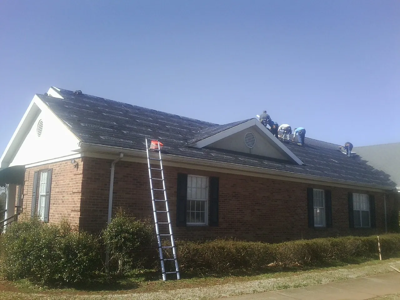 Crew tearing shingles off a brick ranch roof with a ladder set — Emergency Roof Repair work by our crew in Eagle Lake