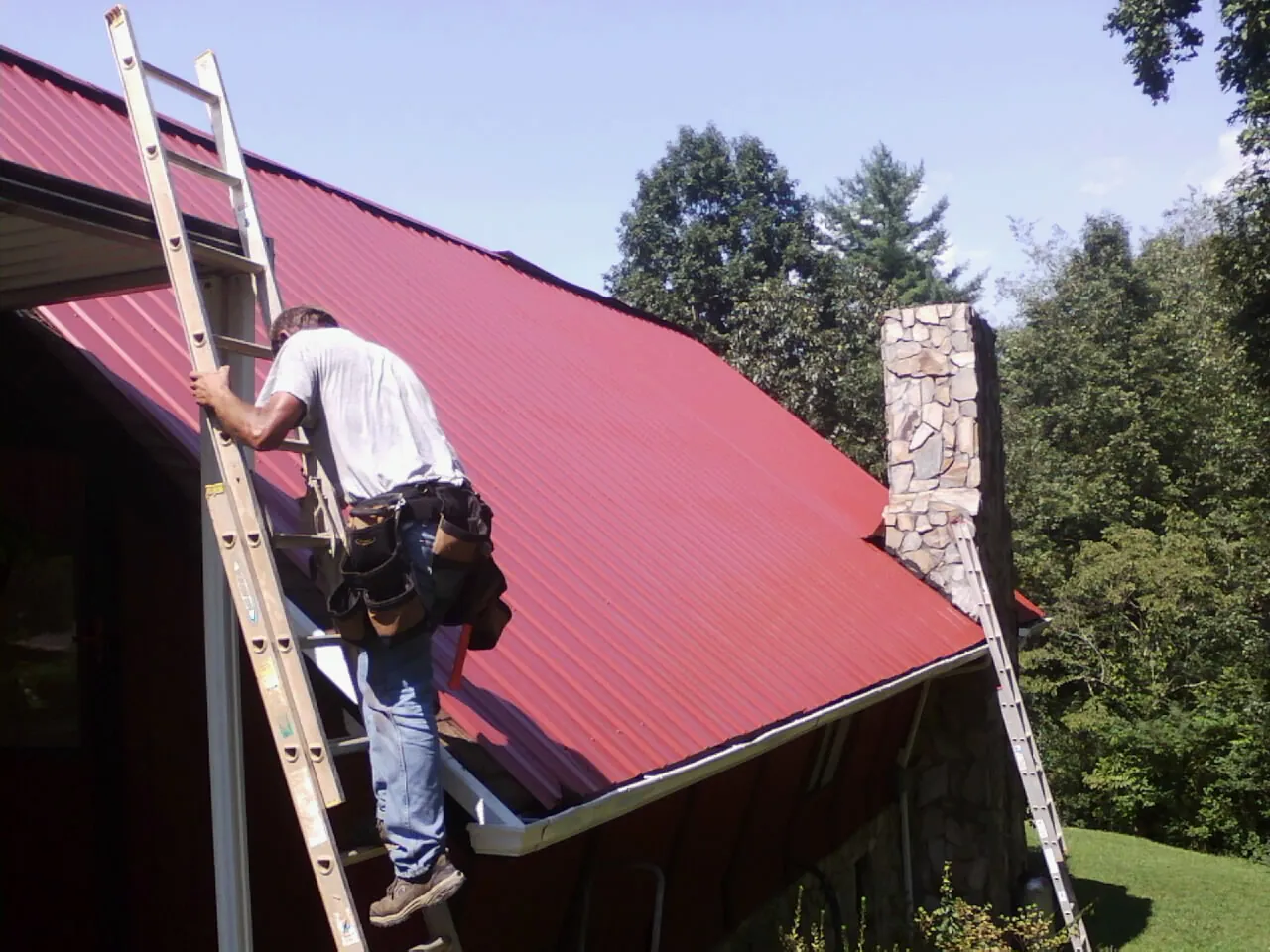 Roofer on a ladder beside a red metal roof and stone chimney — Soffit & Fascia Repair by Roofing Brothers in Eagle Lake